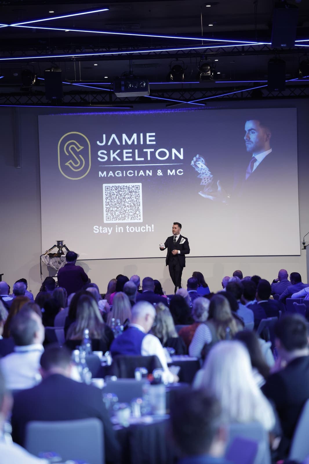 A full conference room at the summit, Jamie small on the stage under a big screen with his name