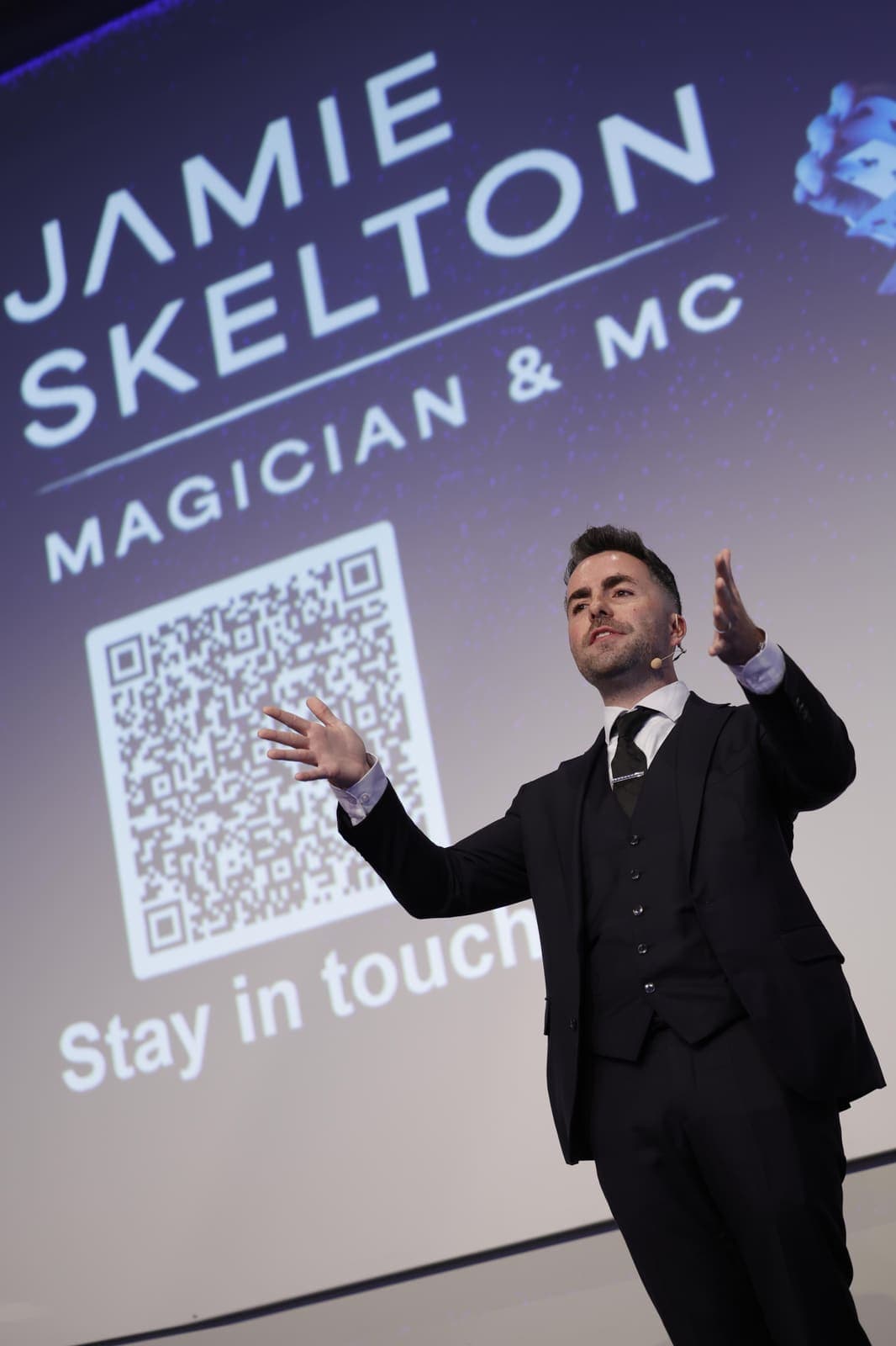 Jamie on the summit stage, arms out, in front of a screen with his name and logo