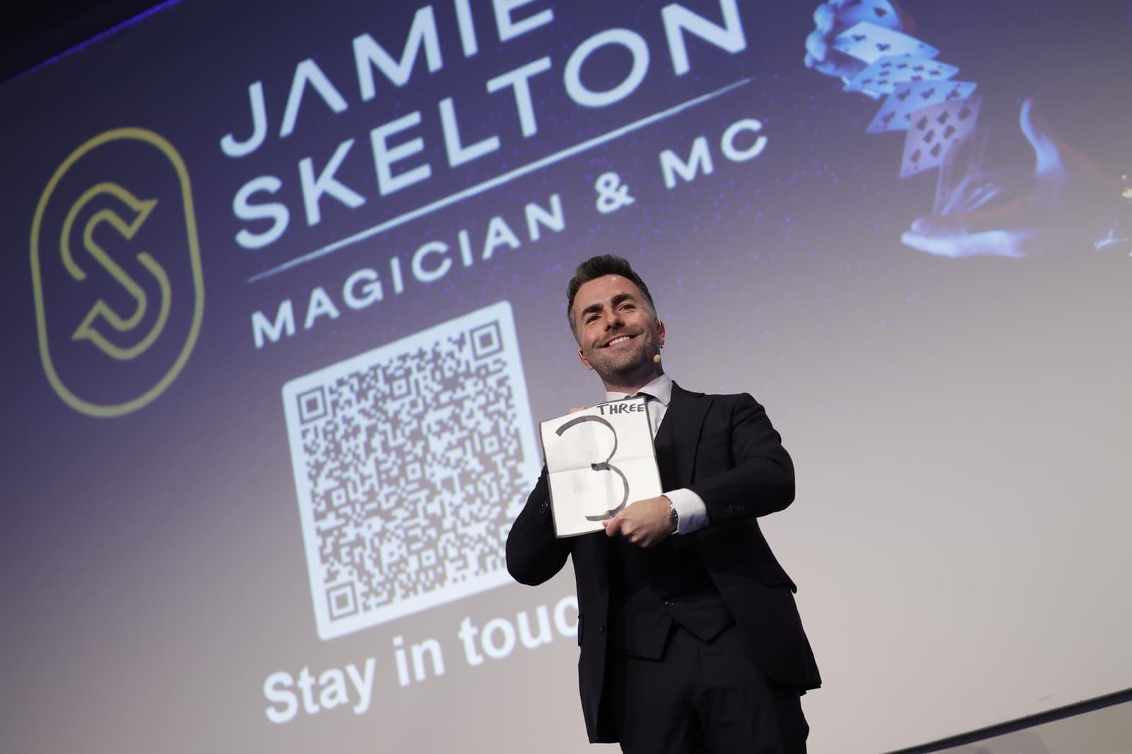 Jamie holds up a card with a drawn number in front of the screen with his logo