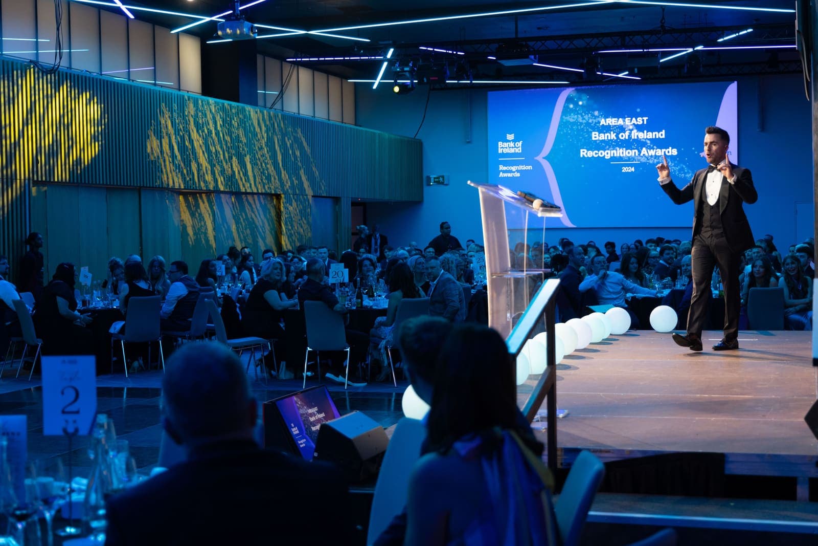 Wide view of the Bank of Ireland Recognition Awards: Jamie on a blue-lit stage above a ballroom of tables