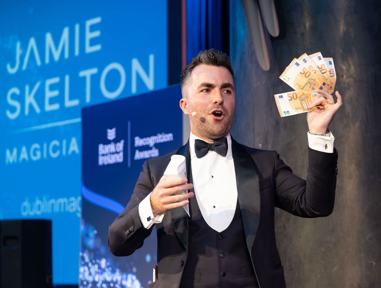 Jamie, with a headset microphone, fans euro banknotes on stage in front of the Bank of Ireland Recognition Awards screens