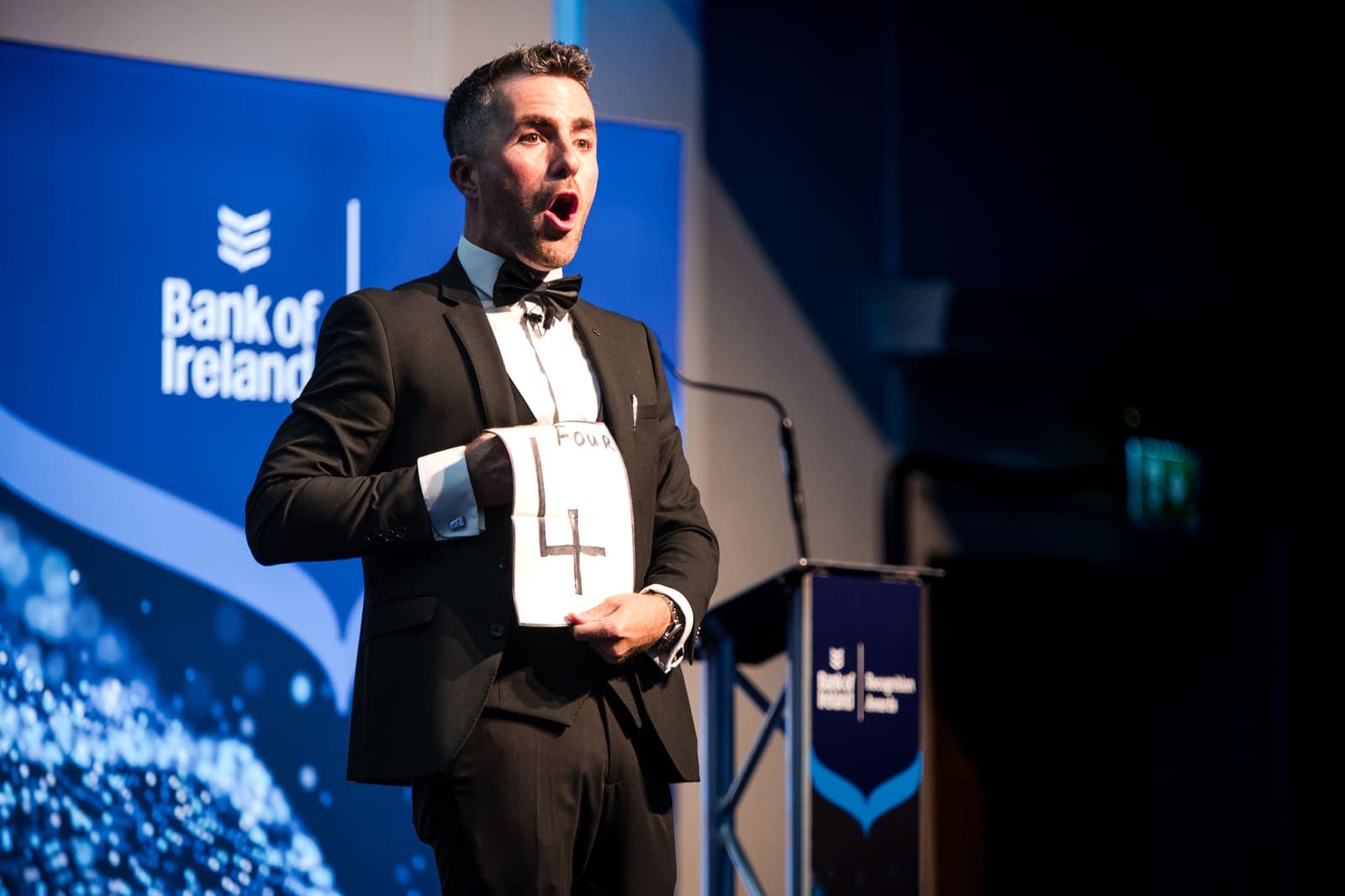 Jamie, mouth open in surprise, holds a card with the number 4 beside the Bank of Ireland lectern