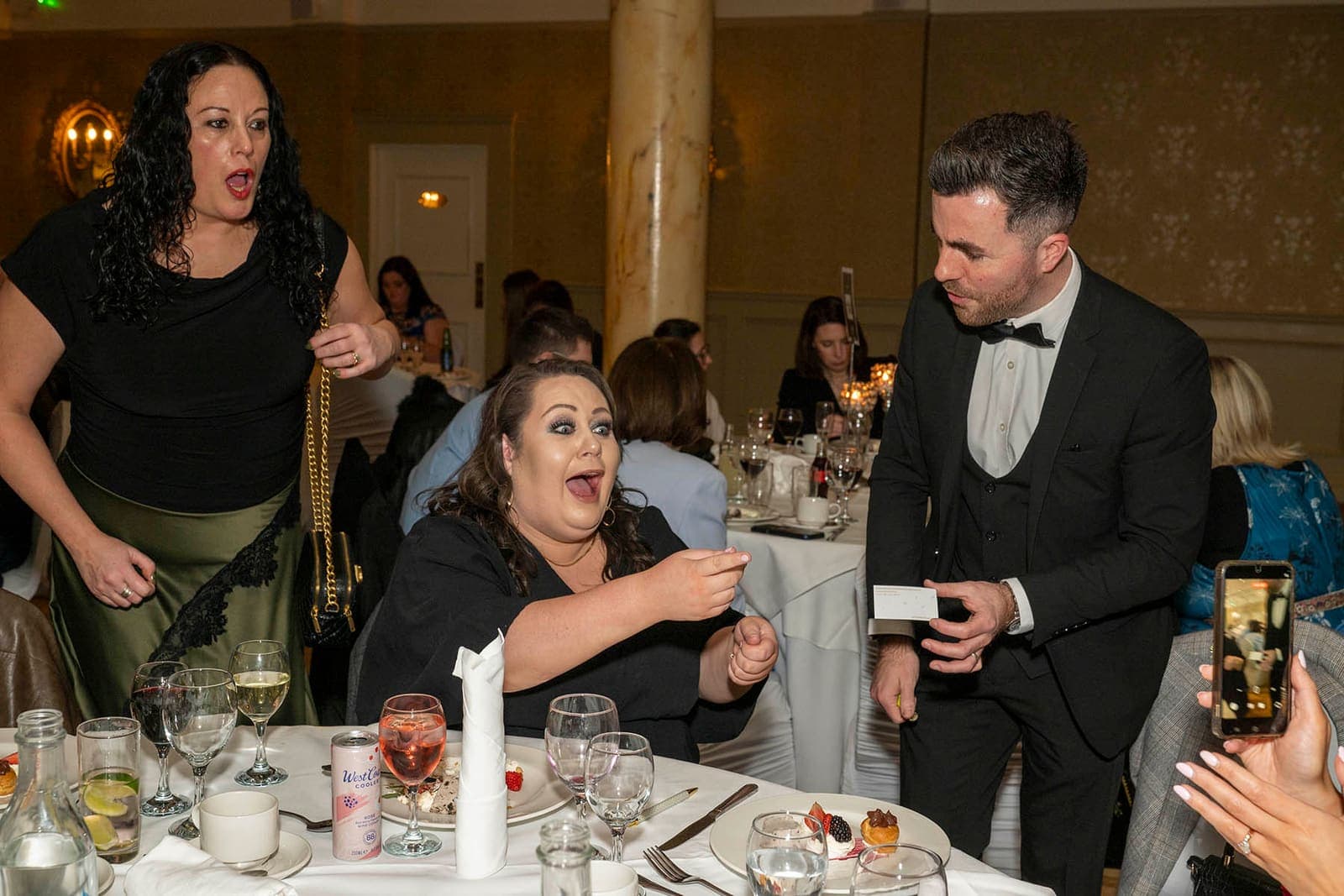 A guest at a dinner table covers her mouth in shock as Jamie shows a card, her friend standing behind her