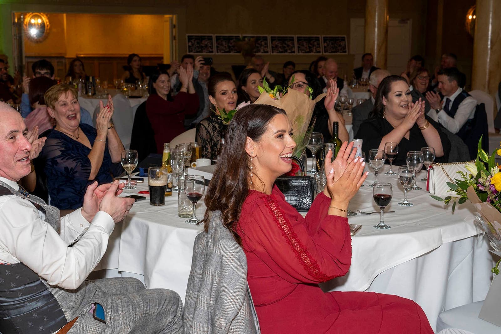 Guests at round tables applaud and laugh, a woman in a red dress in front