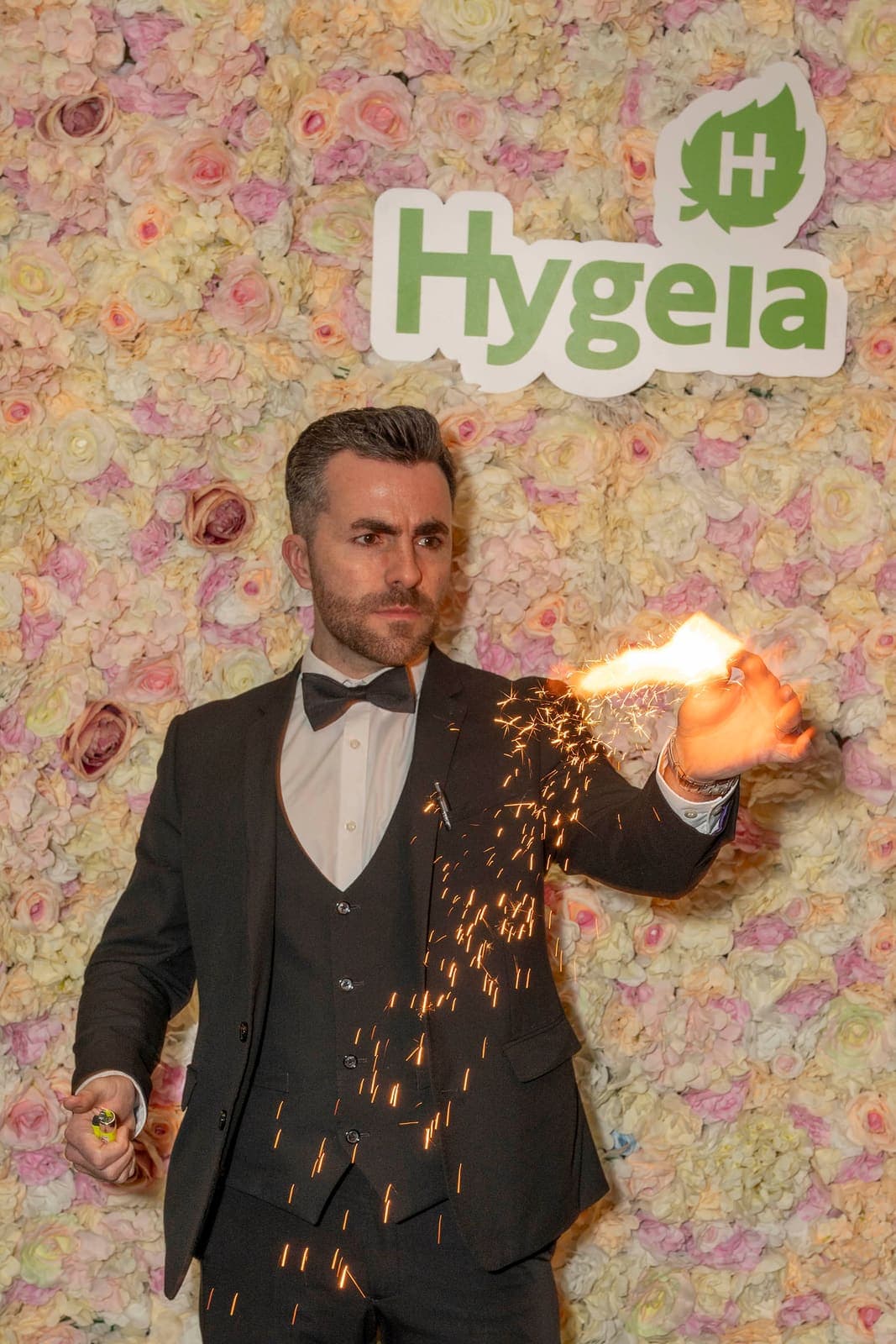 Jamie in a tuxedo throws sparks from his hand in front of the Hygeia flower wall