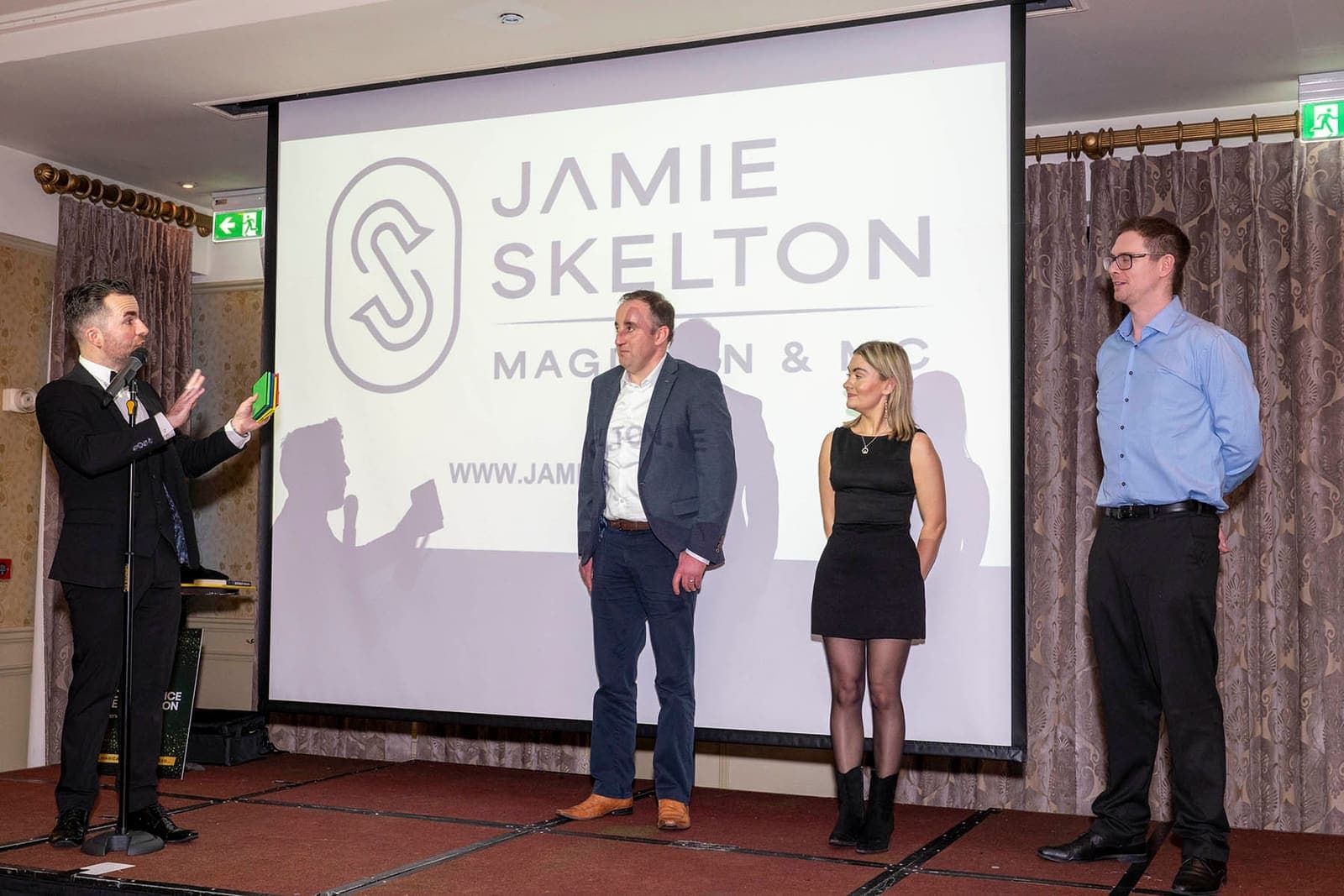 Jamie on stage with three guest volunteers in front of a screen with his logo