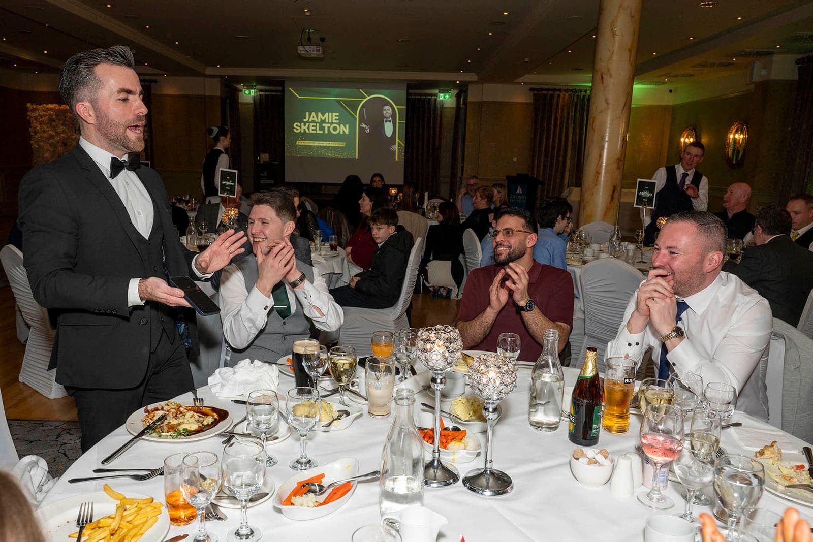 Jamie performs at a dinner table while the guests around it laugh, his logo on the screen at the back of the room