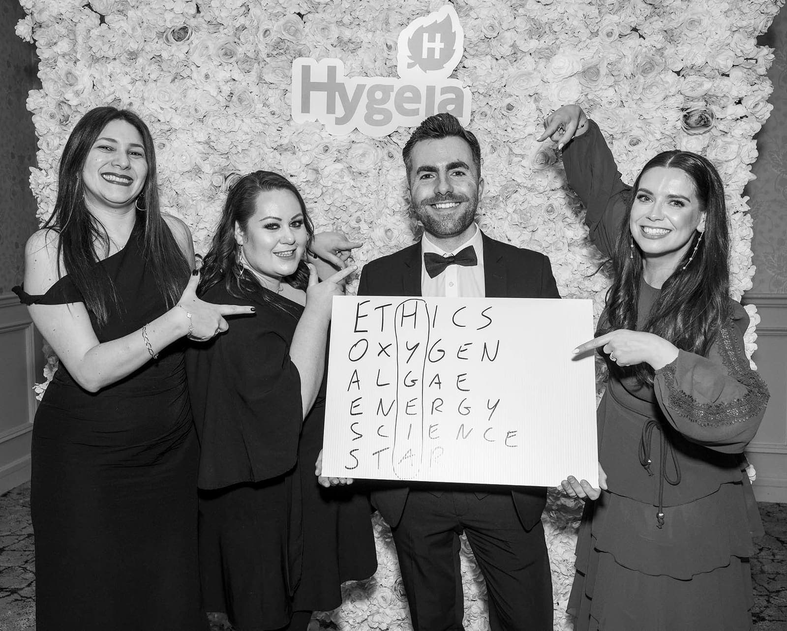 Black and white: Jamie and three guests point at a hand-written word board in front of the Hygeia flower wall