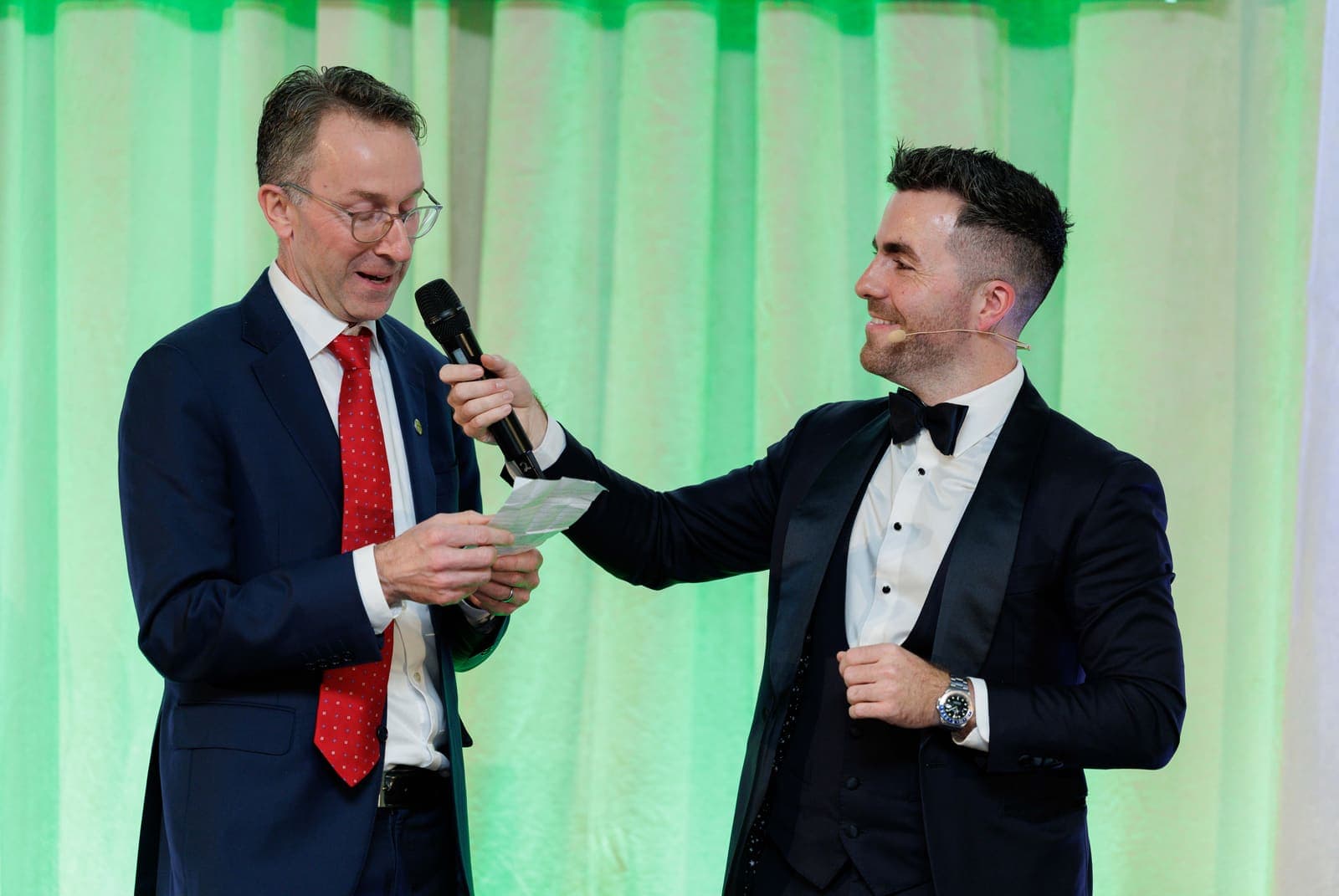 Jamie, in a tuxedo, laughs as a guest in a red tie reads from a card into the microphone