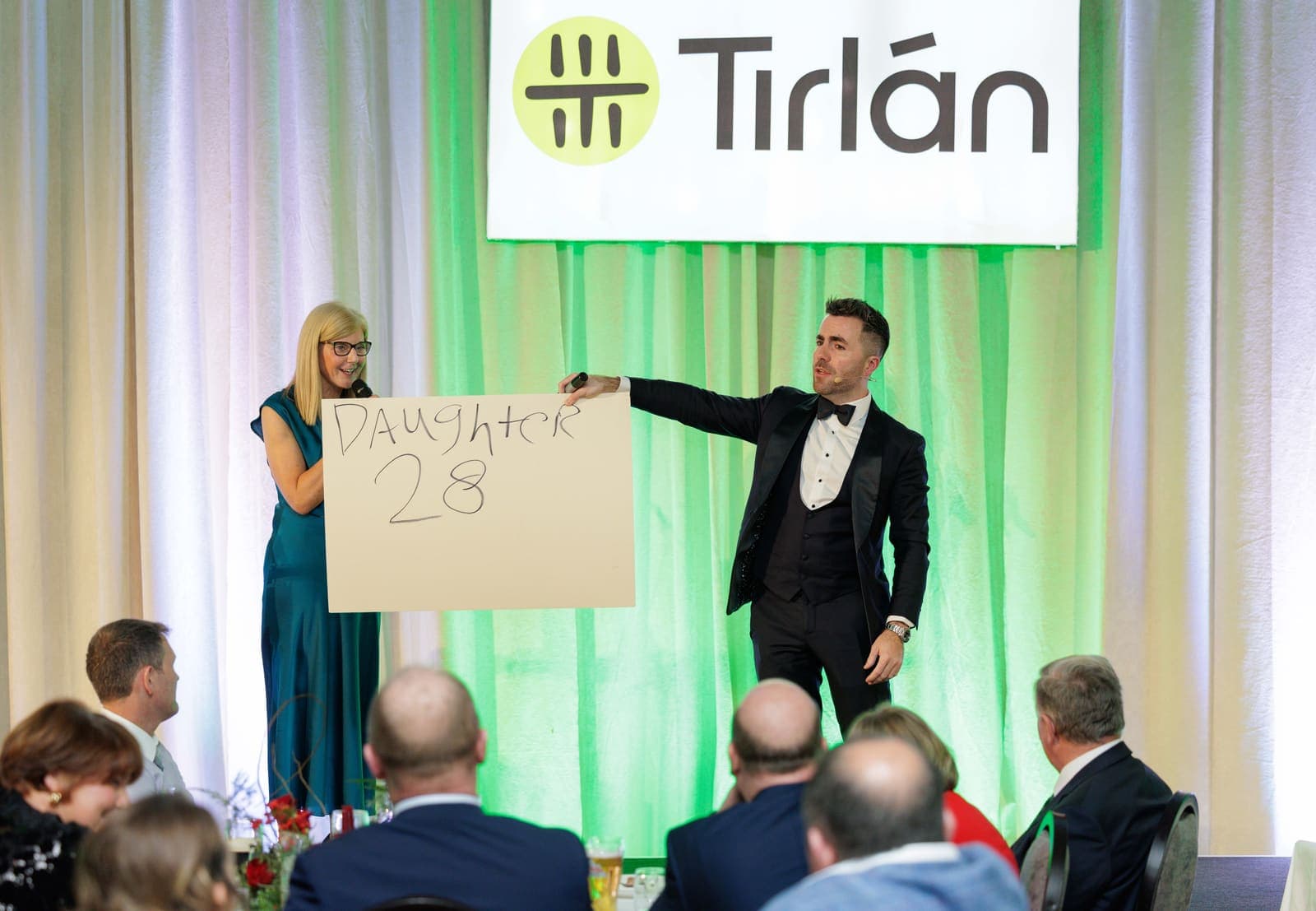 Under a Tirlán sign, Jamie points to a guest holding a hand-written prediction board, the dinner tables in front of the stage