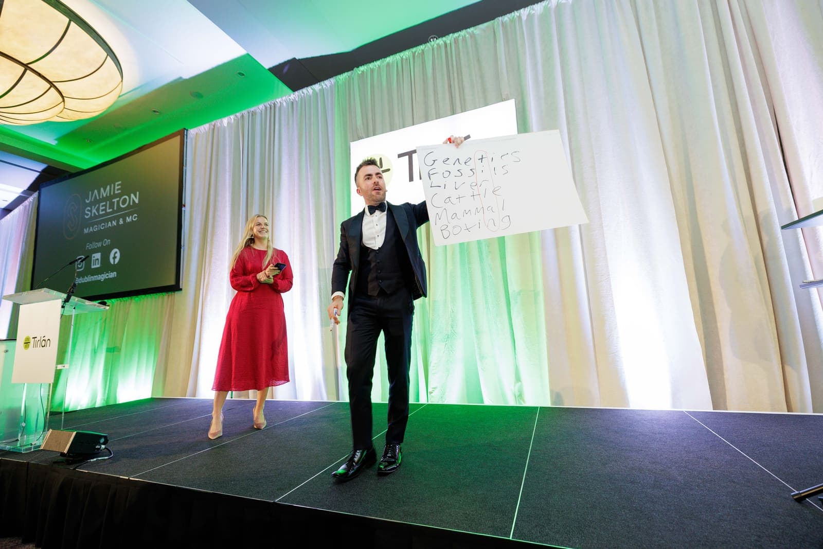 Jamie holds up a large prediction board on stage while a guest in a red dress looks on