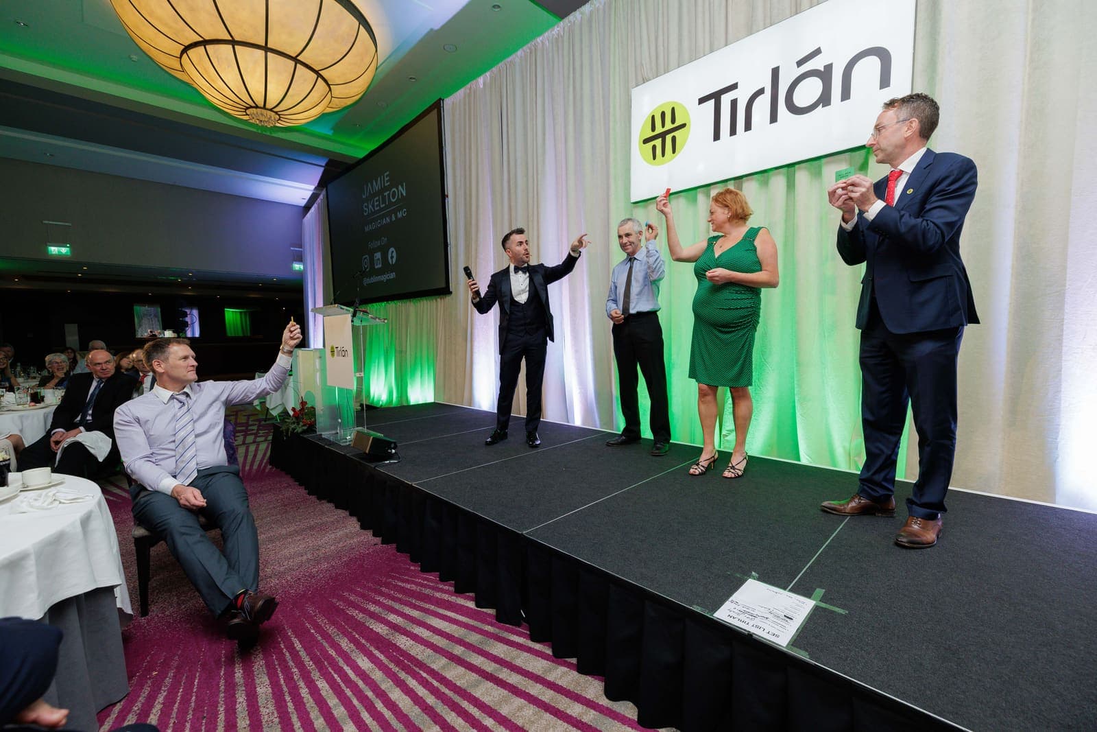 Wide view of the Tirlán council dinner: Jamie on a green-lit stage with three guest volunteers, a guest at a table in front cheering