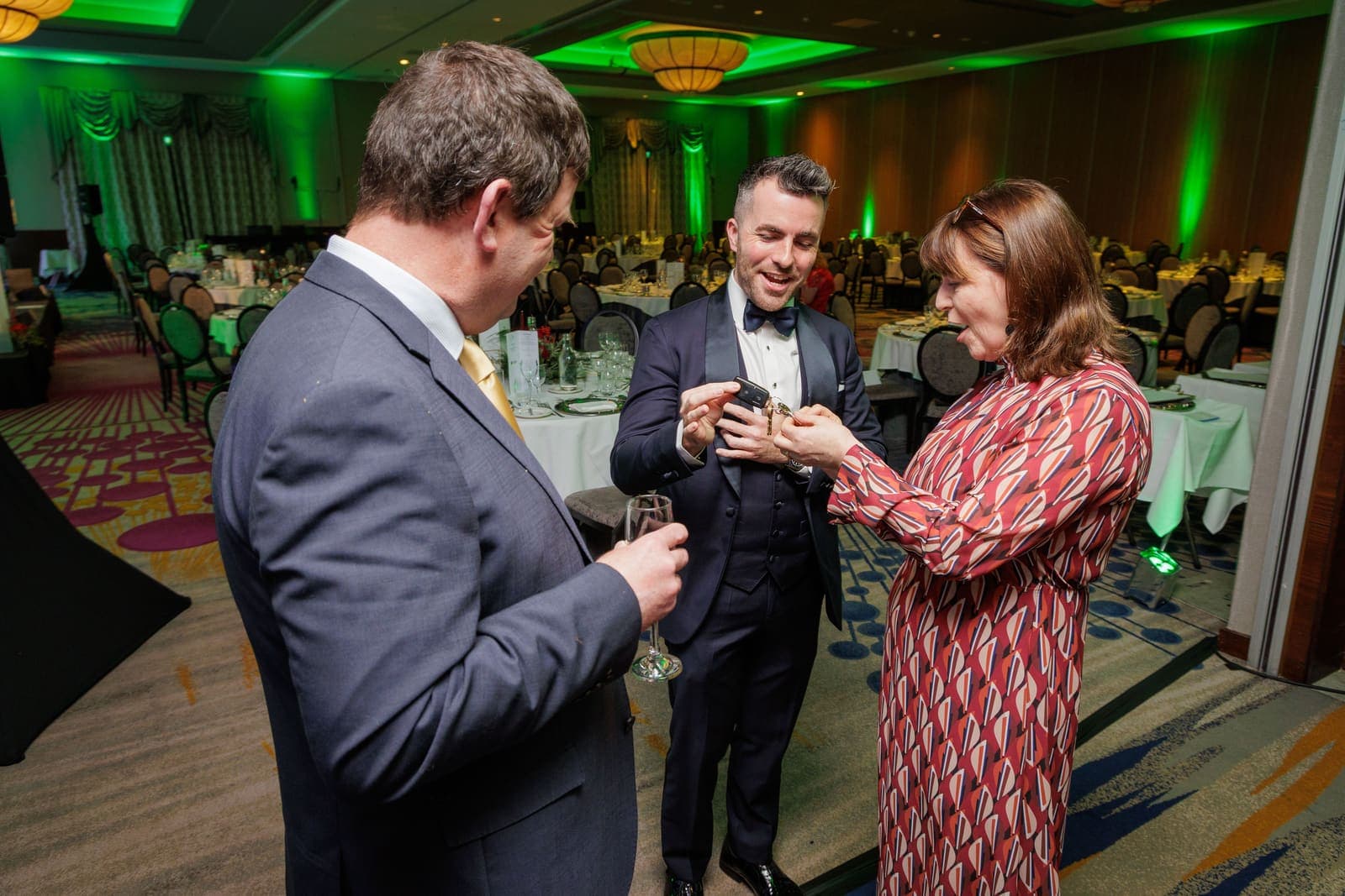 Jamie performs close-up magic for two guests in the green-lit ballroom, one of them smiling at the cards in her hands