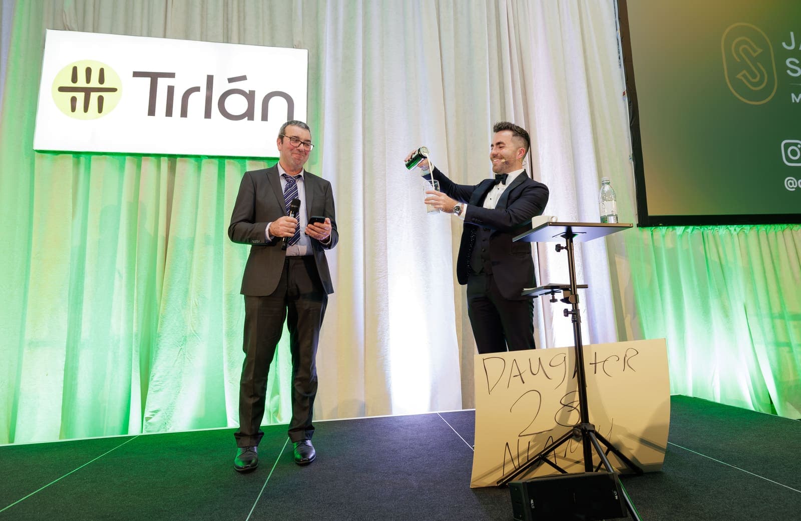 Jamie and a guest volunteer with a microphone on the Tirlán stage, a prediction board on a stand beside them