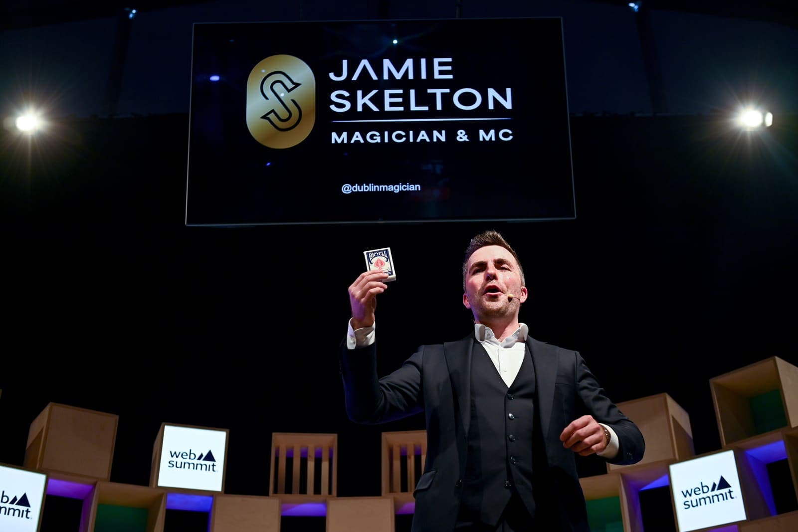 Jamie holds up a card box on the dark Web Summit stage, his gold logo on the screen above