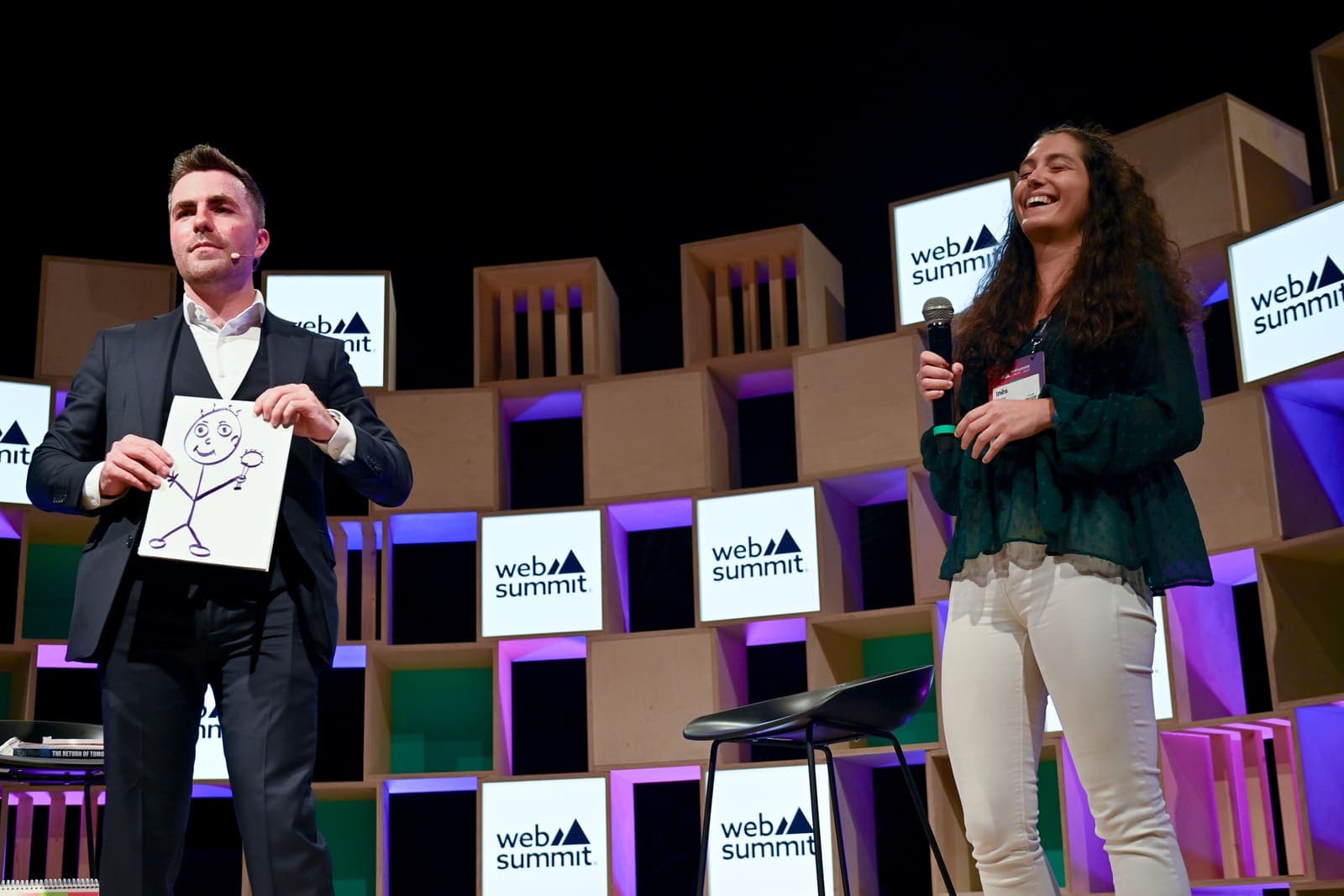 Jamie shows his drawing beside a laughing volunteer holding a microphone on the Web Summit stage