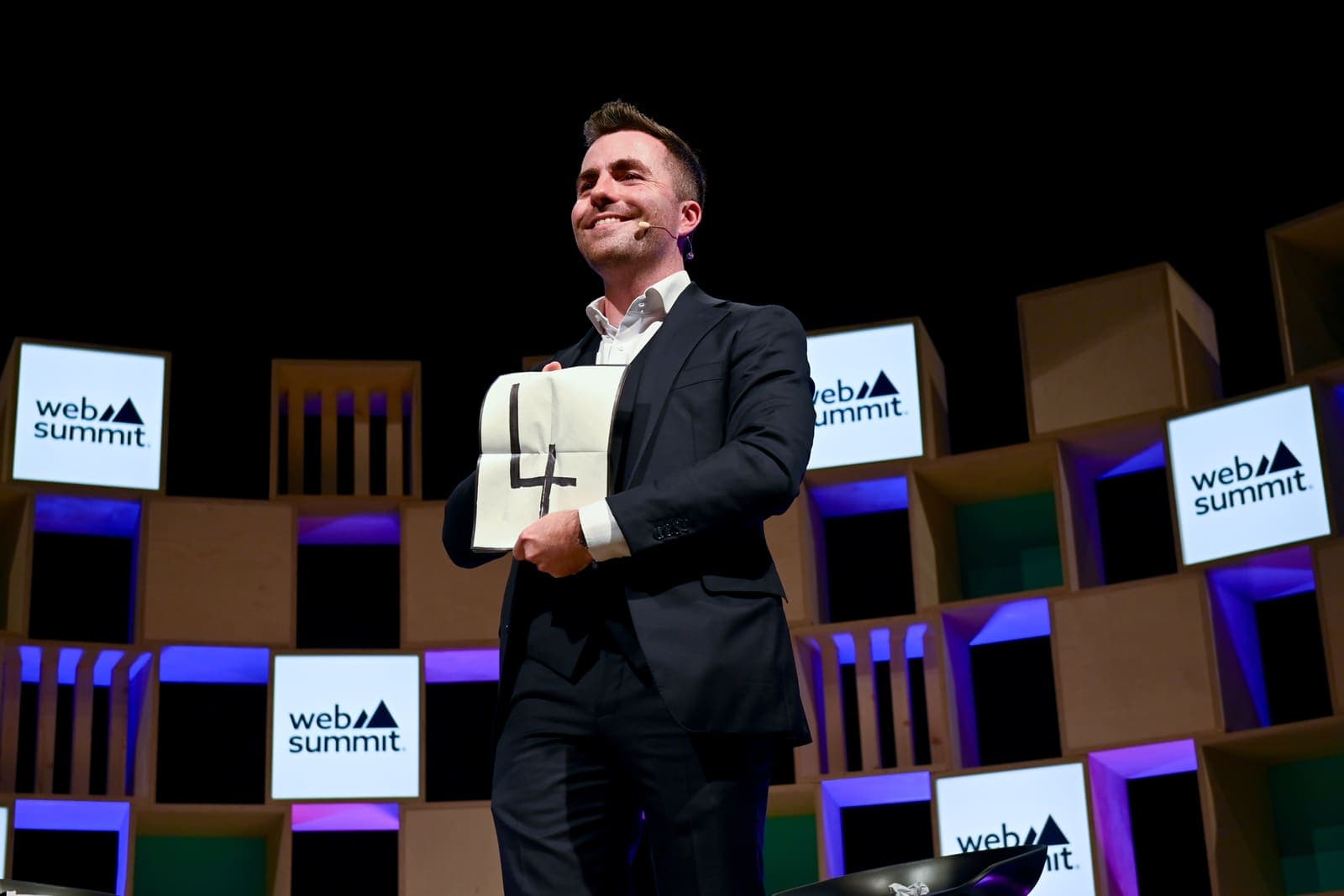 Jamie with a headset microphone holds a large prediction card with a drawn number on the Web Summit stage