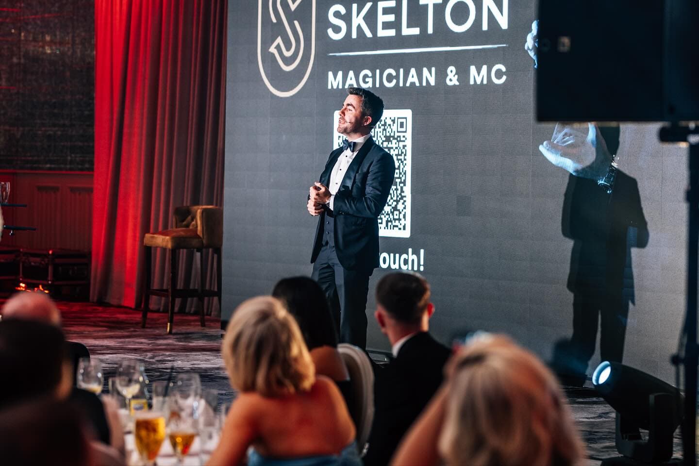 Jamie in a tuxedo on stage at the Wedding Help Northern Ireland Awards 2025, under a screen with his name, guests at their tables in front