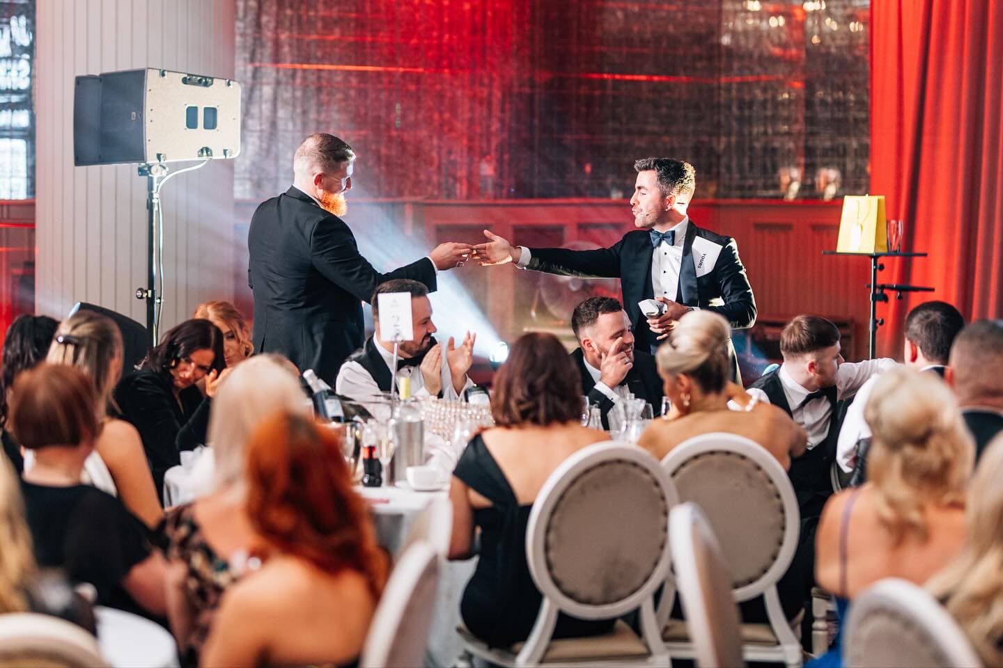 Jamie hands a card to a bearded guest above the tables at the Wedding Help Northern Ireland Awards 2025 as the table claps