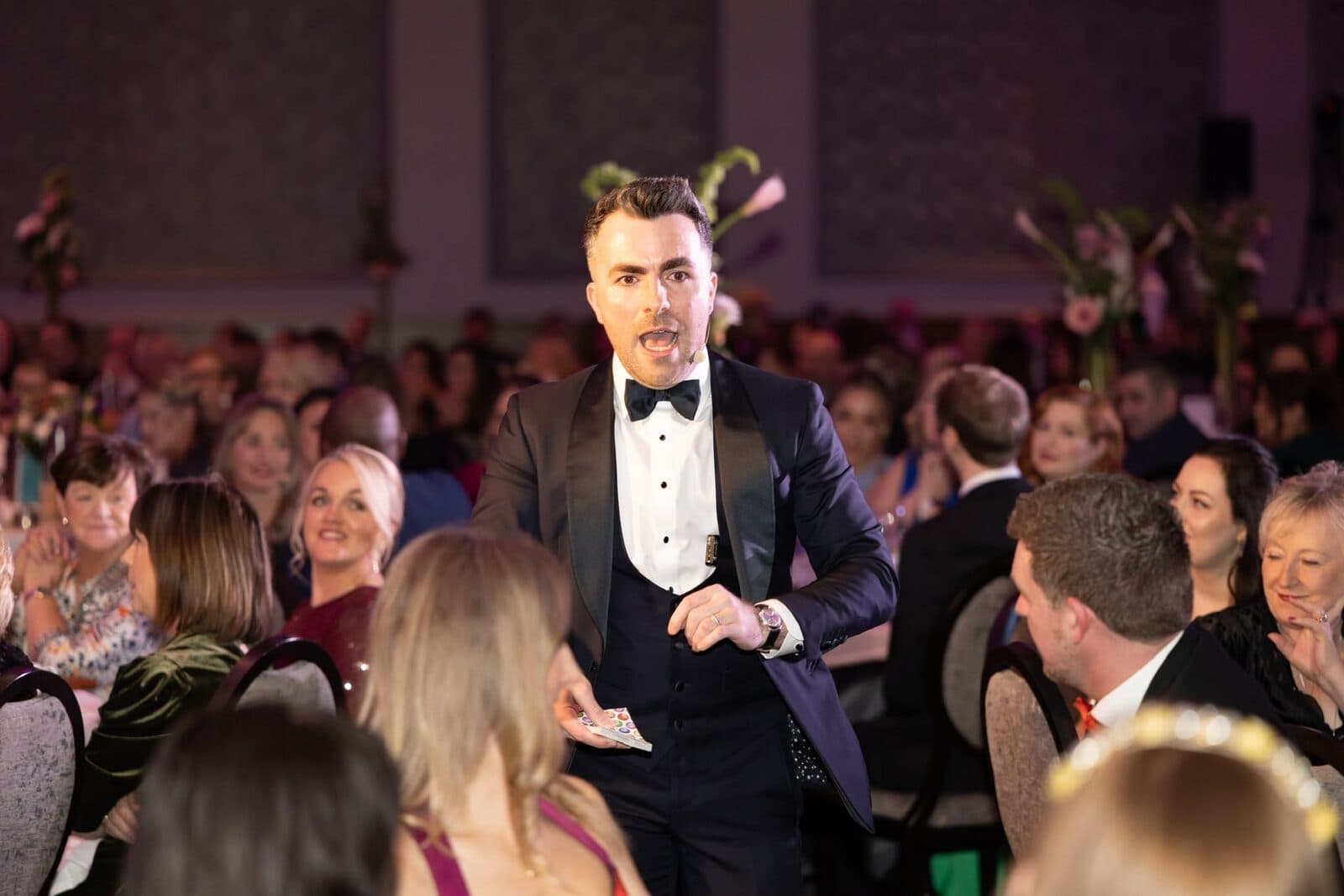 Jamie in a tuxedo and headset microphone acts amazed among the tables at the weddingsonline Awards 2025 as guests smile