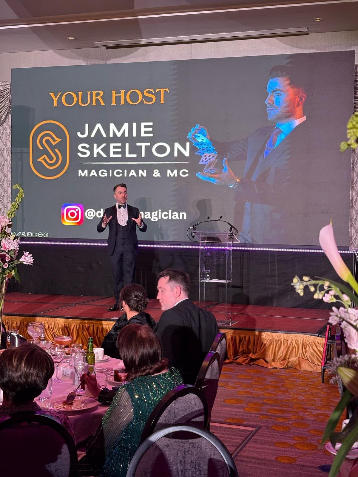 Jamie talks to the room from the weddingsonline Awards 2025 stage under a big screen that reads Your Host, Jamie Skelton, Magician & MC