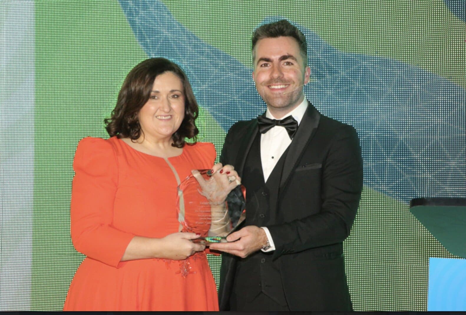 Jamie in a tuxedo presents a glass award to a woman in orange on a stage lit green and blue