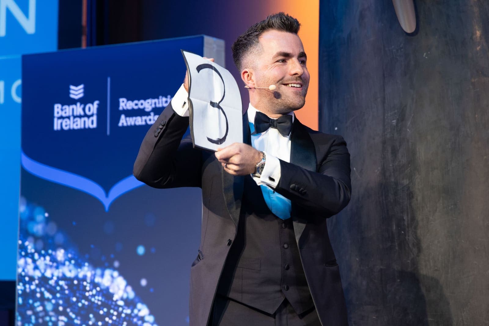Jamie, wearing a headset microphone, smiles as he holds up a number card on stage at the Bank of Ireland Recognition Awards