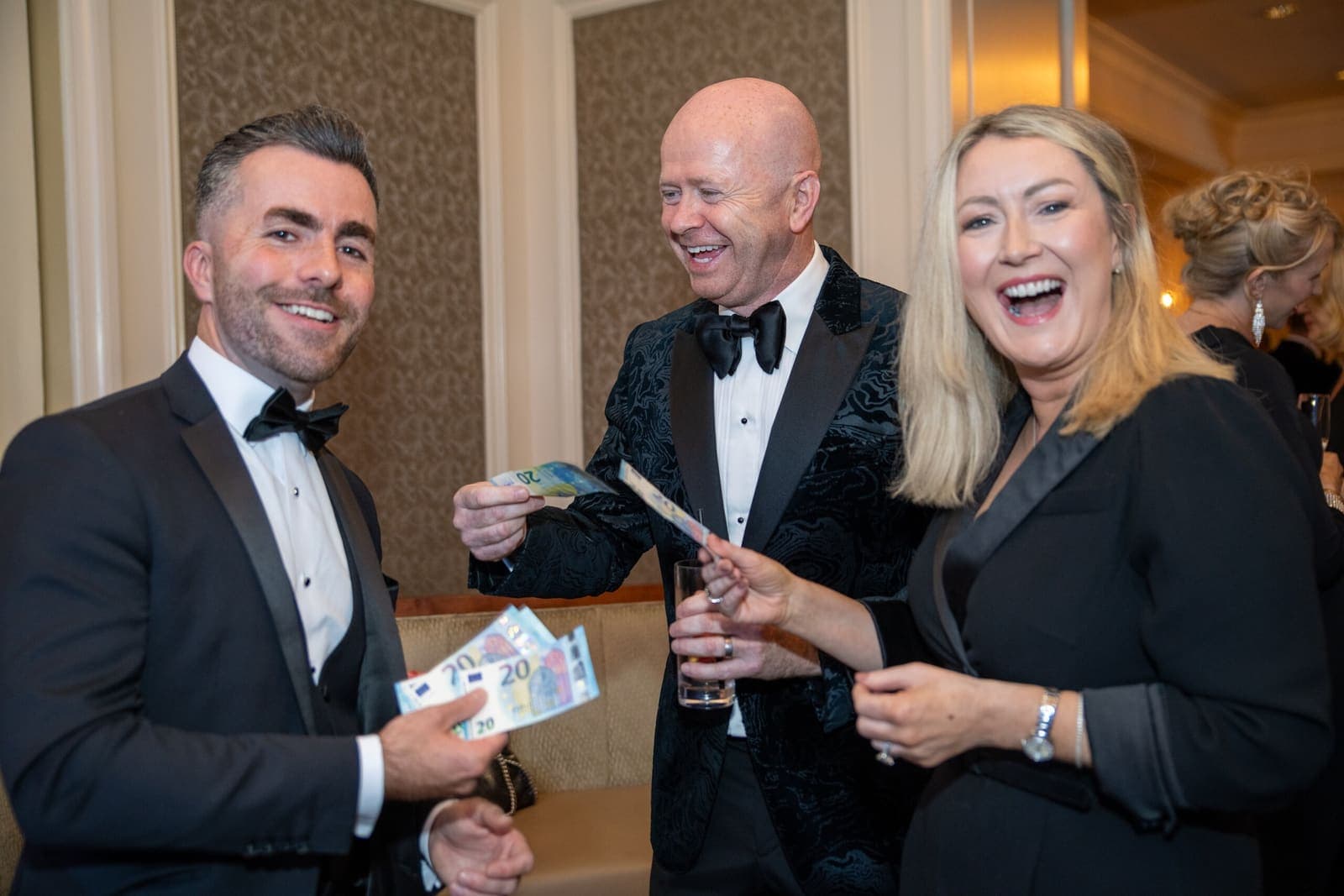 Jamie in black tie holds a fan of €20 notes; a man and a woman beside him laugh, each with a note