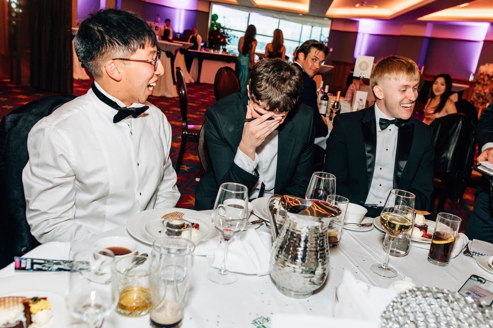 Young men in black tie cover their faces in disbelief at a gala