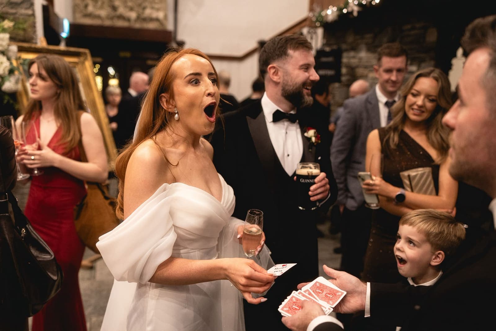A bride gasps at a card trick while a young guest beside her stares open-mouthed