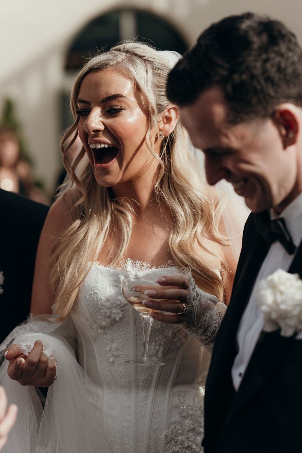 A bride gasps open-mouthed as the groom beside her grins at a trick
