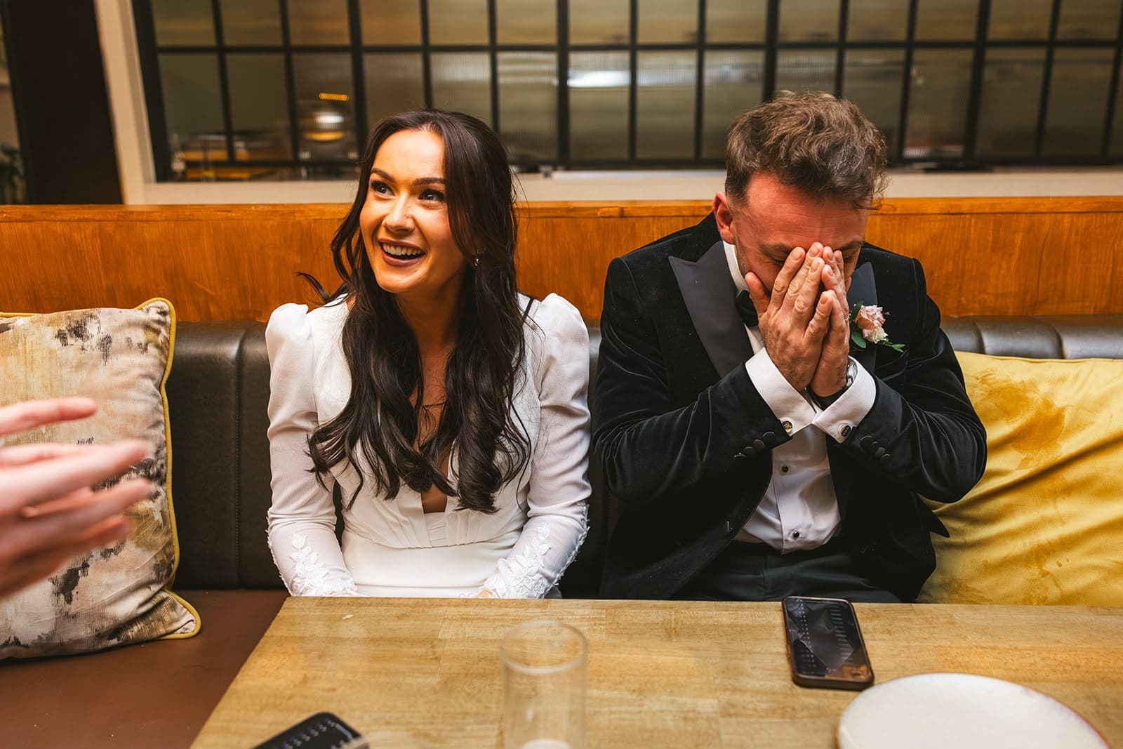 A bride laughs while the groom beside her hides his face in his hands