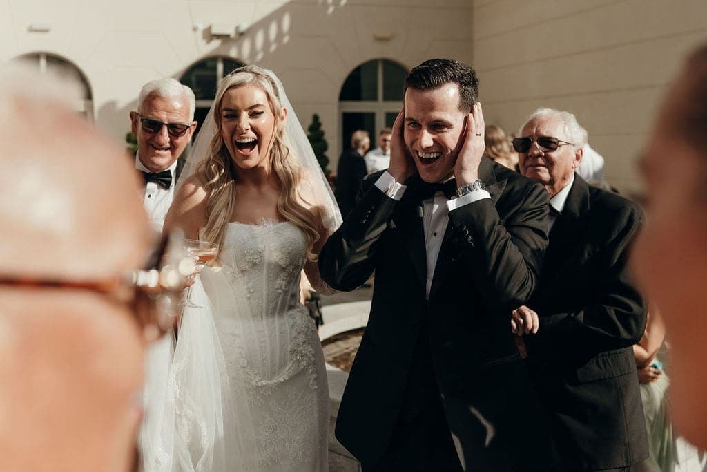 A bride laughs and the groom grabs his head in disbelief, with older guests behind them