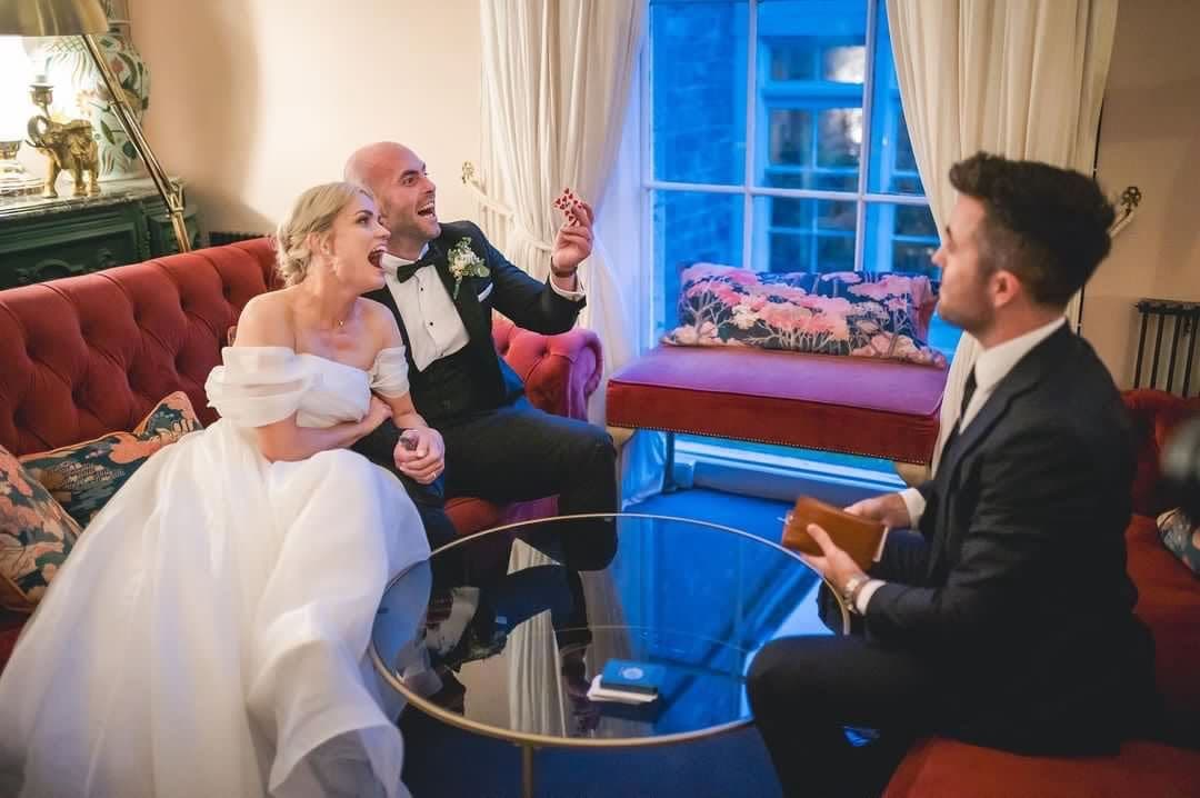 A bride and groom on a red sofa laugh as Jamie shows them a trick