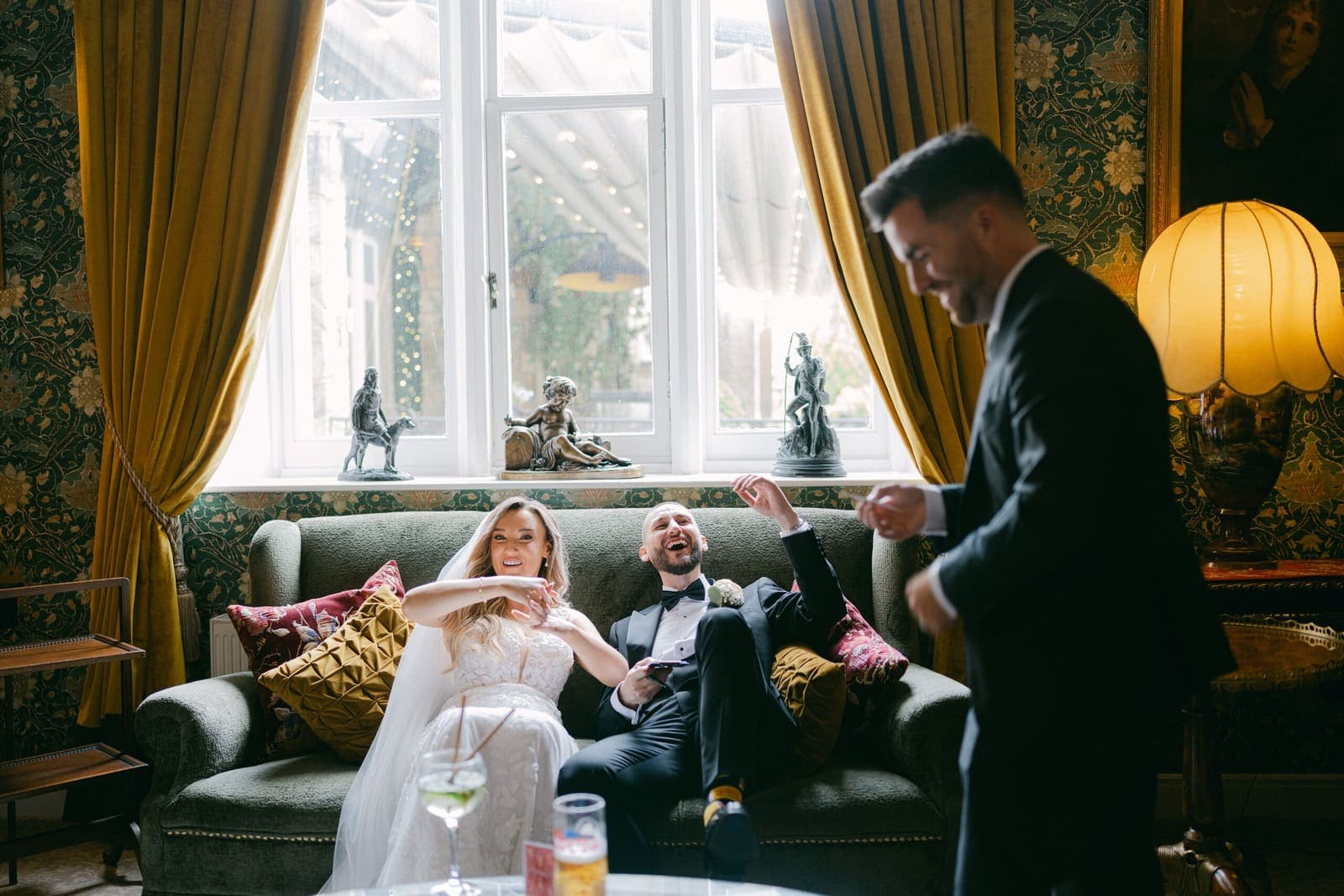 Bride and groom on a velvet sofa in an ornate drawing room watch Jamie