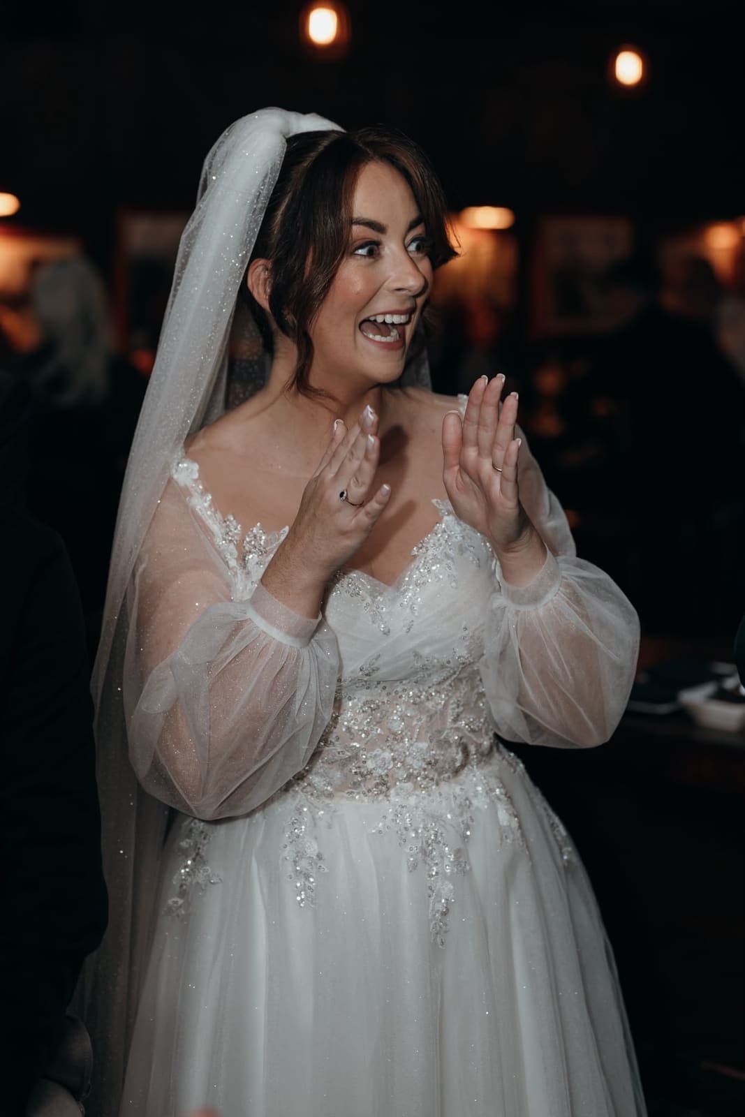 A bride in her veil gasps and raises her hands, wide-eyed, at a trick