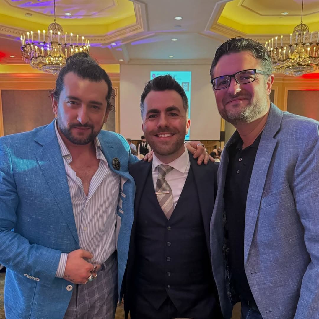 Jamie smiles between two men in blazers in a ballroom with chandeliers