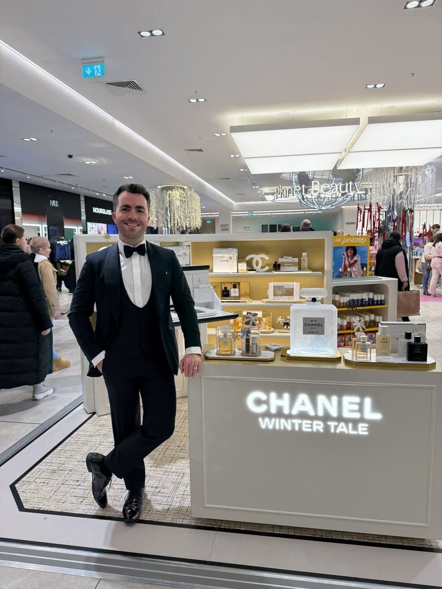 Jamie in black tie leans on a lit Chanel Winter Tale counter in a department store