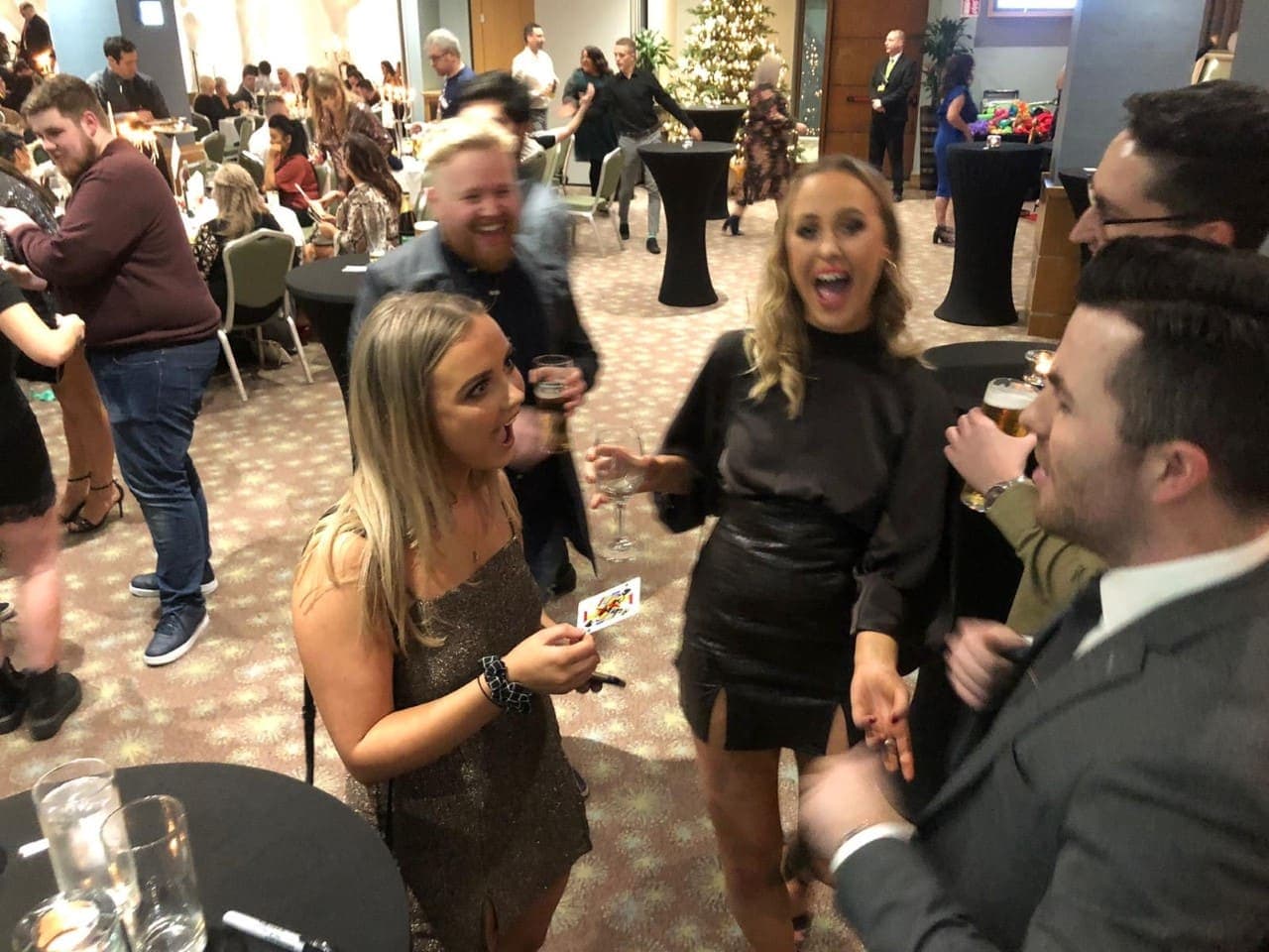 At a party with a Christmas tree behind, a woman in a black dress laughs with her mouth open beside a friend who holds a playing card
