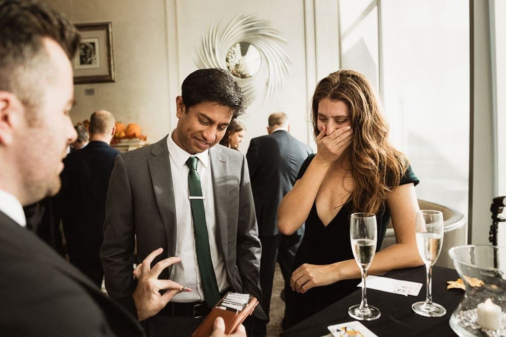 At a cocktail table, a guest covers her mouth and laughs at a card trick