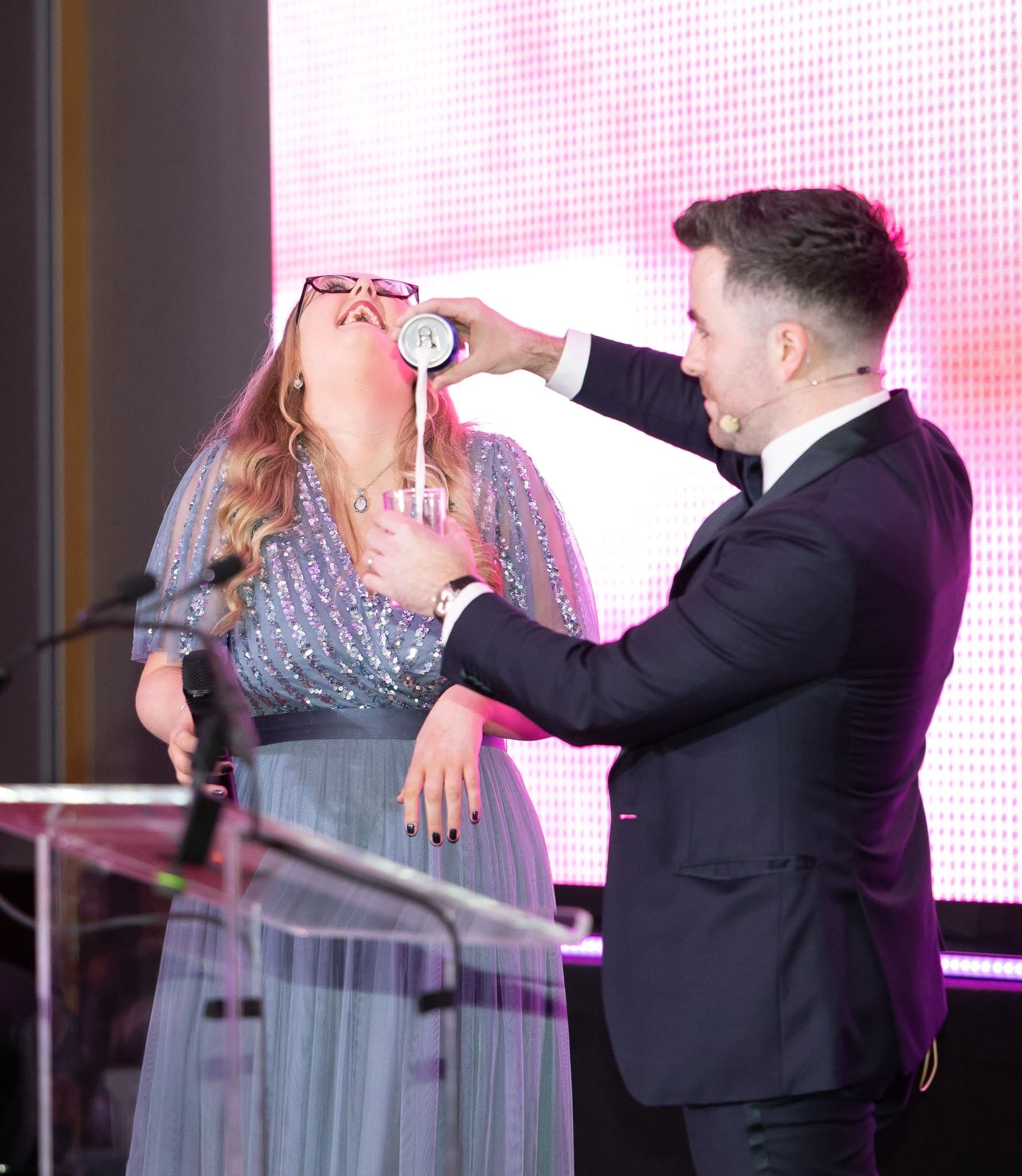 On a stage with an LED screen and a lectern, Jamie, wearing a headset microphone, pours a drink for a laughing volunteer