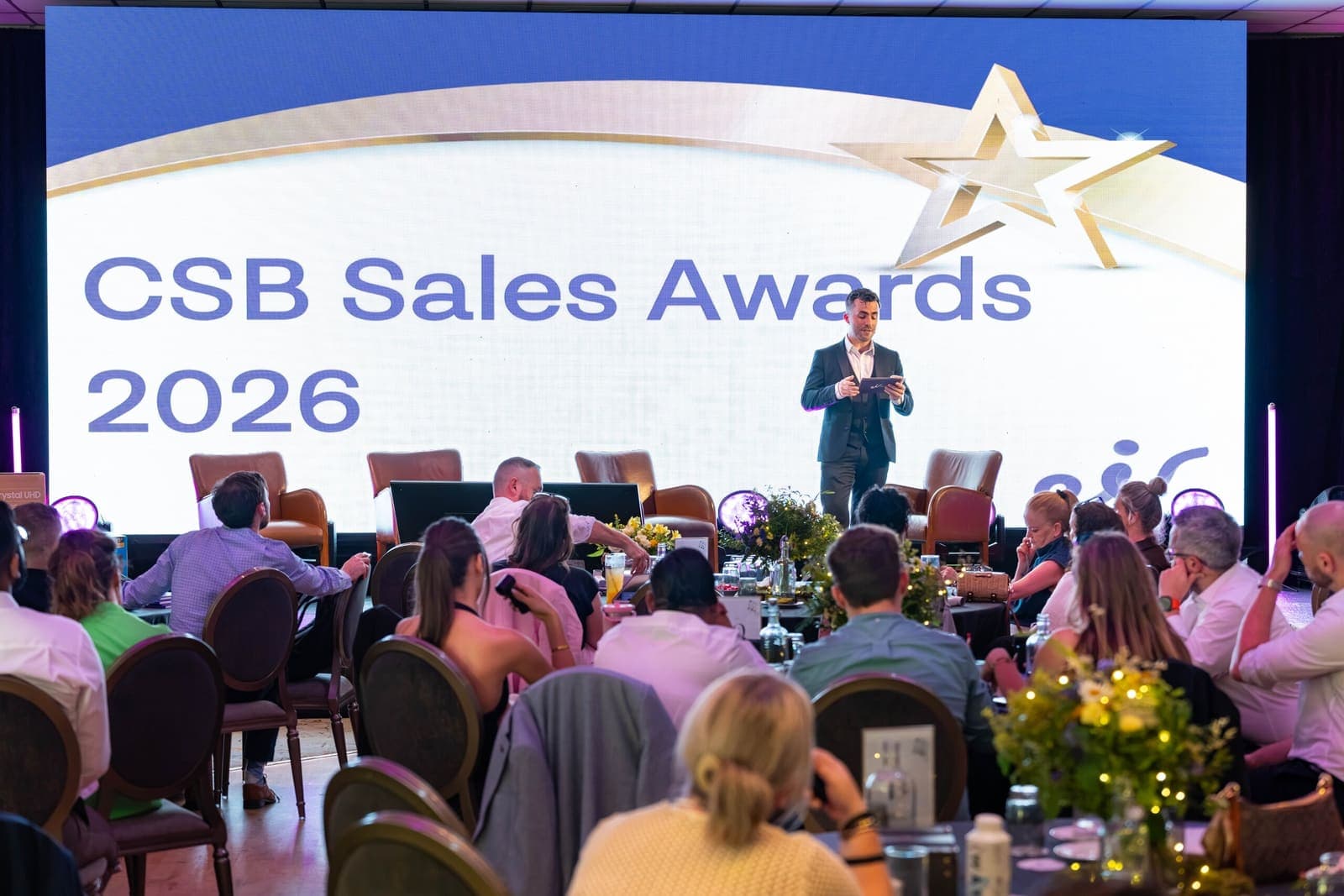 Jamie in a suit reads a card on stage under a big CSB Sales Awards 2026 screen, guests at tables in front