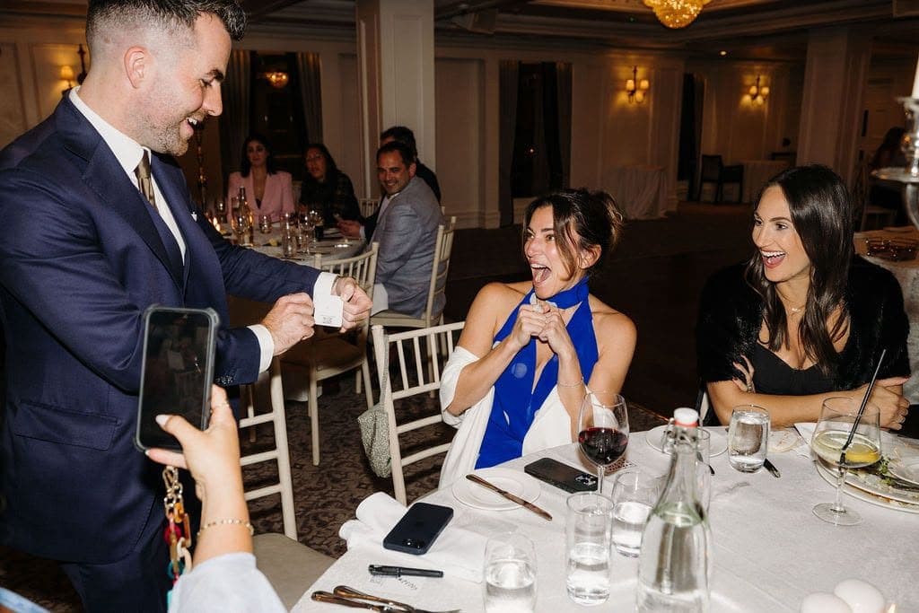 At a dinner table, a guest clasps her hands and gasps with delight as Jamie finishes a trick; her friend laughs