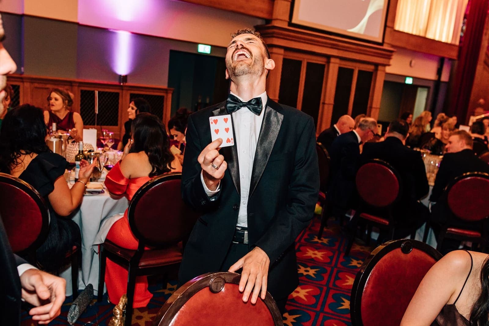 Jamie laughs while showing a card at a formal dinner in a red-carpeted ballroom