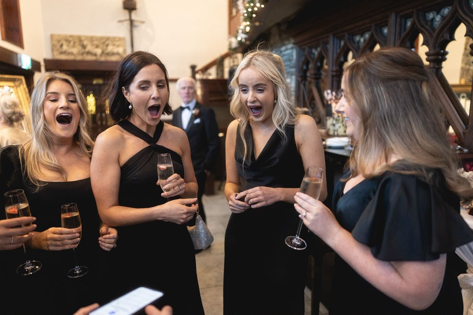Friends in black dresses, glasses in hand, gasp with open mouths at a trick
