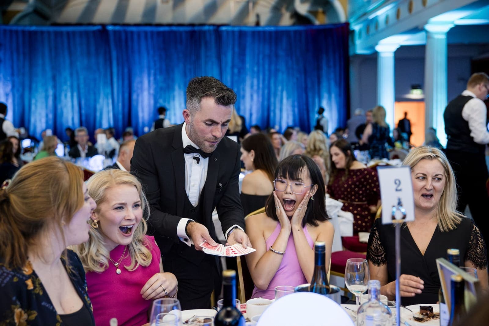 At a gala dinner table, two guests gasp, one with both hands on her cheeks, as Jamie fans out the cards