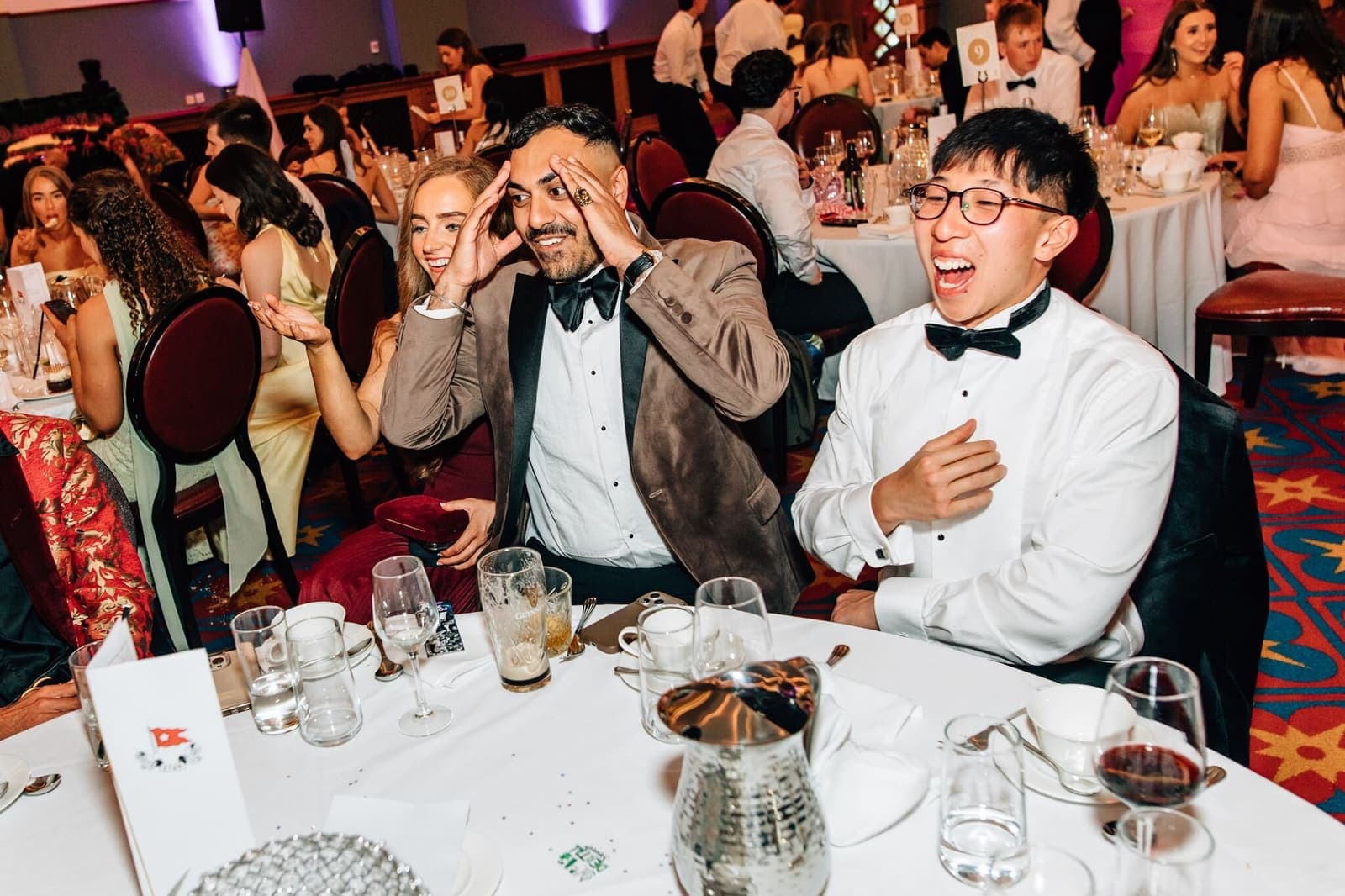 Two men in black tie at a gala dinner table hold their heads, stunned by a trick