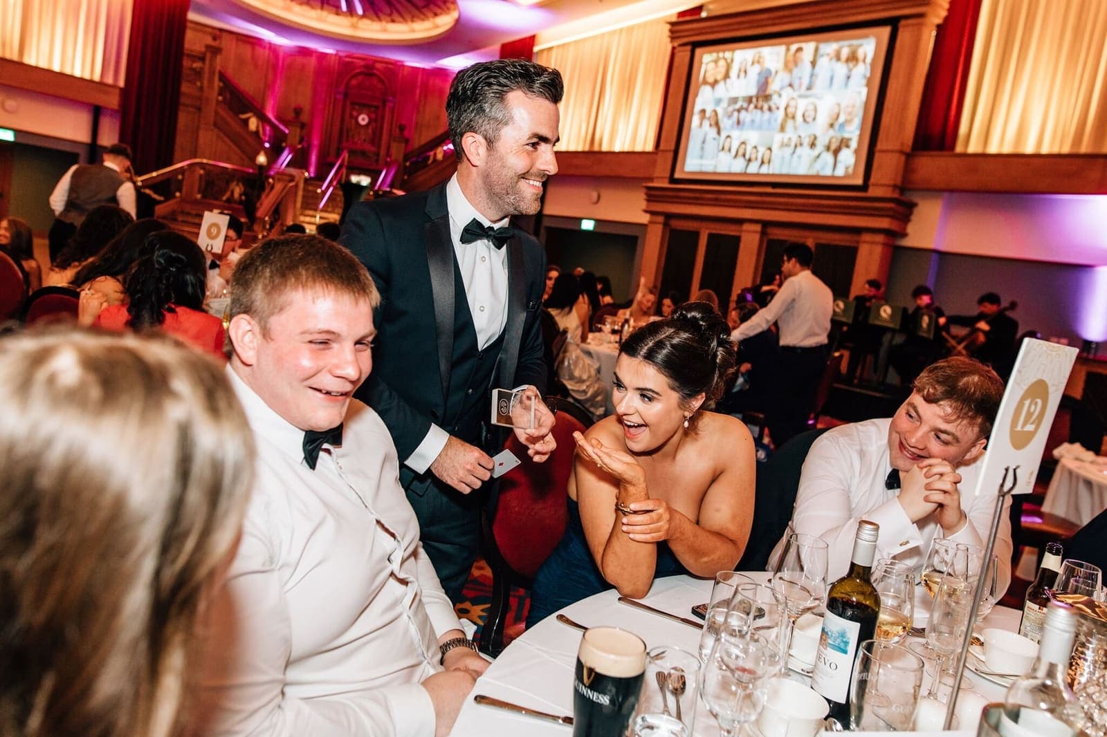Jamie in a tuxedo performs at a round table in a red and gold ballroom; guests in black tie laugh