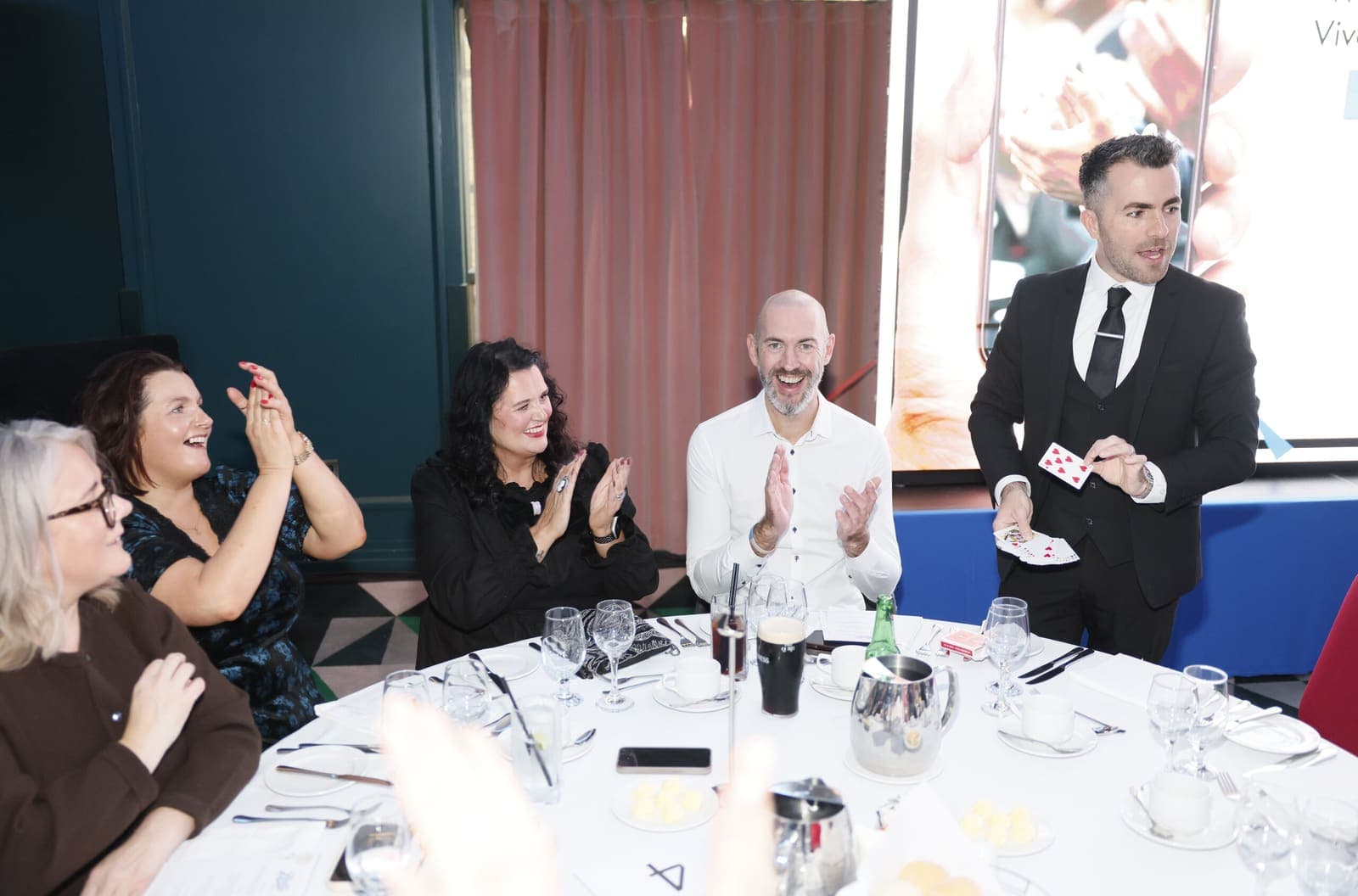 Jamie in a suit shows a card at a dinner table; guests clap and laugh
