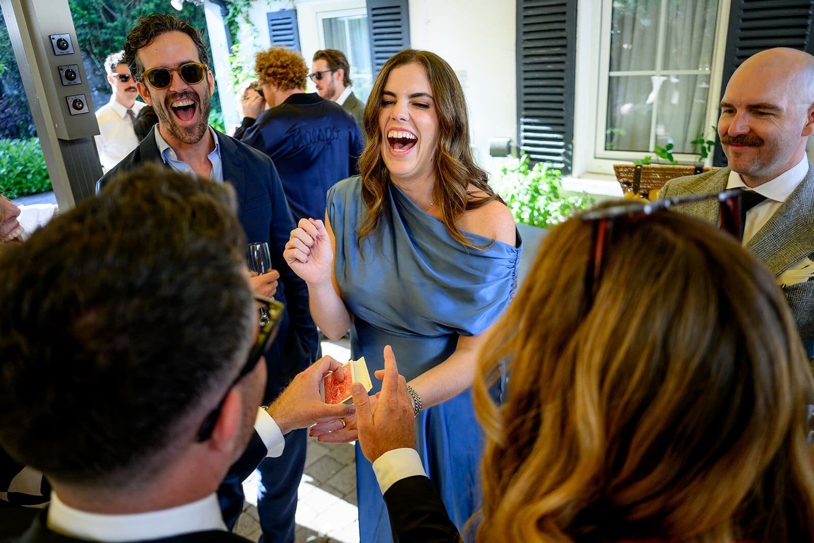 At a garden drinks reception, two wedding guests burst out laughing, close to the camera, as Jamie finishes a card trick
